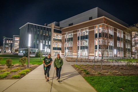 Two people walk along a lit sidewalk at night in front of MSU's STEM building with glass windows and bright interior lighting, surrounded by landscaped grounds and campus lighting.
