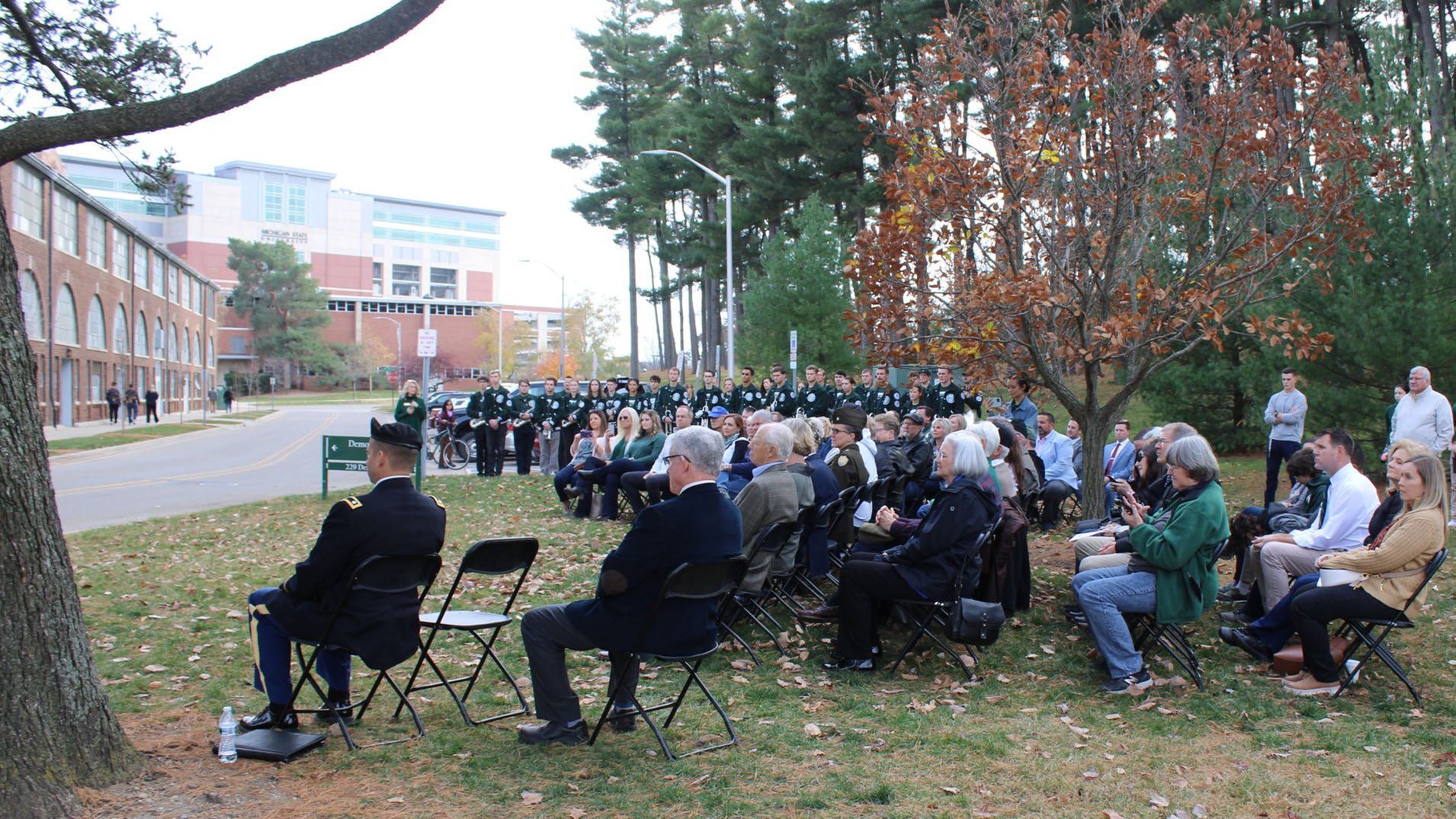 attendees sit in chairs on the lawn across from the plaza location and listen to the presentation