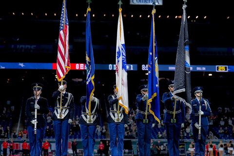 Military color guard in a stadium setting holding various flags including the Michigan state flag and US flag. Camera view is facing toward them and seven people standing in a line. 