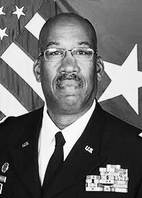 Black and white portrait photo. Subject is seen from chest up, wearing a military uniform, with visible medals on their shirt. They are standing against an American flag.