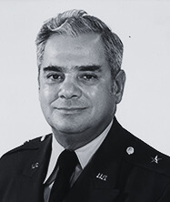 Black and white portrait photo. Subject is seen from shoulders up with visible medals on their military uniform. They are against a white backdrop. 