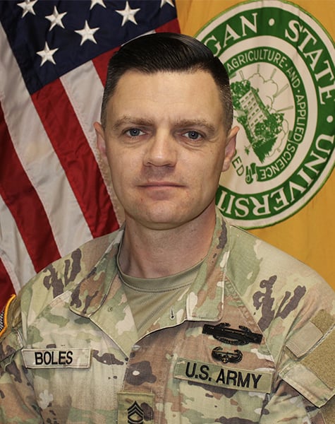 Portrait image. Subject is seen from mid-chest up and are looking at the camera with minimal expression. They are wearing Army fatigues and sitting in front of an American flag and yellow flag with Michigan State University seal on it.