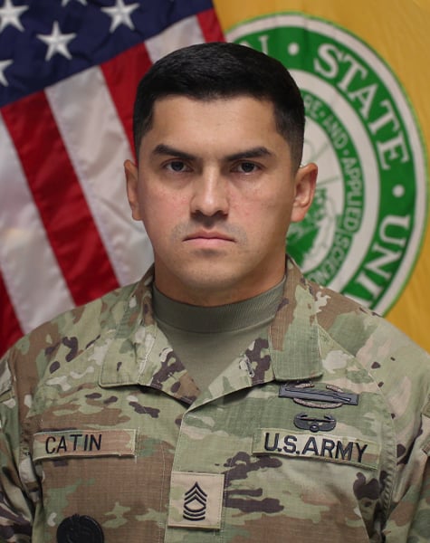 Portrait image. Subject is seen from mid-chest up and are looking at the camera with minimal expression. They are wearing Army fatigues and sitting in front of an American flag and yellow flag with Michigan State University seal on it.