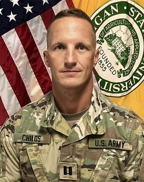 Portrait image. Subject is seen from mid-chest up and are looking at the camera with minimal expression. They are wearing Army fatigues and sitting in front of an American flag and yellow flag with Michigan State University seal on it.