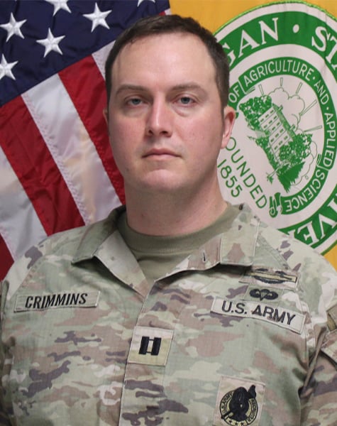 Portrait image. Subject is seen from mid-chest up and are looking at the camera with minimal expression. They are wearing Army fatigues and sitting in front of an American flag and yellow flag with Michigan State University seal on it. 