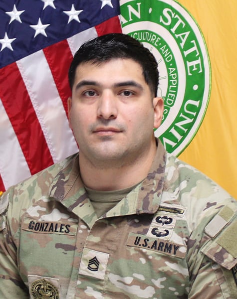 Portrait image. Subject is seen from mid-chest up and are looking at the camera with minimal expression. They are wearing Army fatigues with patches reading "US Army" on one side and "Gonzales" on the other. They are sitting in front of an American flag and yellow flag with Michigan State University seal on it.