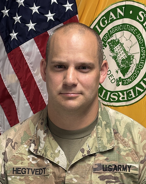 Portrait image. Subject is seen from mid-chest up and are looking at the camera with minimal expression. They are wearing Army fatigues and sitting in front of an American flag and yellow flag with Michigan State University seal on it. 