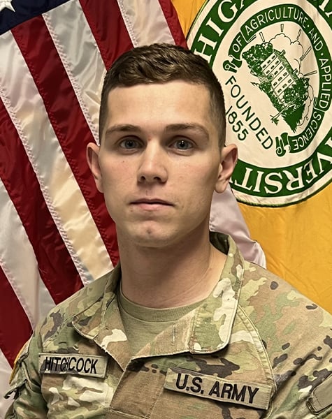 Portrait image. Subject is seen from mid-chest up and are looking at the camera with minimal expression. They are wearing Army fatigues and sitting in front of an American flag and yellow flag with Michigan State University seal on it.