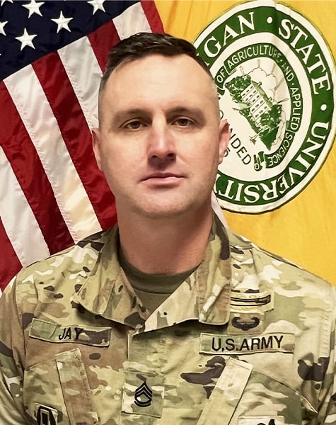 Portrait image. Subject is seen from mid-chest up and are looking at the camera with minimal expression. They are wearing Army fatigues and sitting in front of an American flag and yellow flag with Michigan State University seal on it.