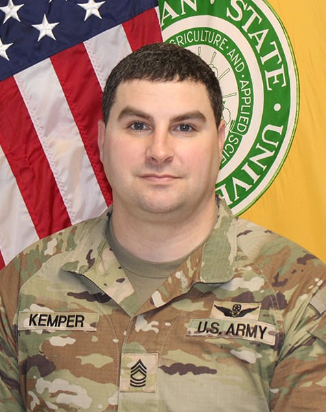 Portrait image. Subject is seen from mid-chest up and are looking at the camera with minimal expression with their dark hair cut short. They are wearing Army fatigues and sitting in front of an American flag and yellow flag with Michigan State University seal on it.