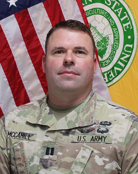 Portrait image. Subject is seen from mid-chest up and are looking at the camera with minimal expression. They are wearing Army fatigues and sitting in front of an American flag and yellow flag with Michigan State University seal on it.
