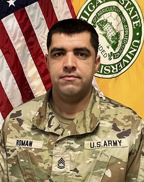 Portrait image. Subject is seen from mid-chest up and are looking at the camera with minimal expression. They are wearing Army fatigues and sitting in front of an American flag and yellow flag with Michigan State University seal on it.