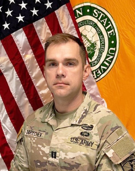 Portrait. Subject is featured from mid-chest up, standing in front of an American flag and yellow flag. They are wearing Army fatigues with hair cut short. 