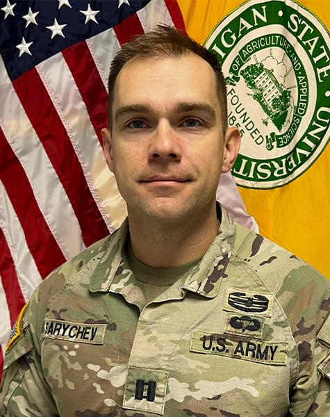 Portrait image. Subject is seen from mid-chest up and are looking at the camera with minimal expression. They are wearing Army fatigues and sitting in front of an American flag and yellow flag with Michigan State University seal on it.