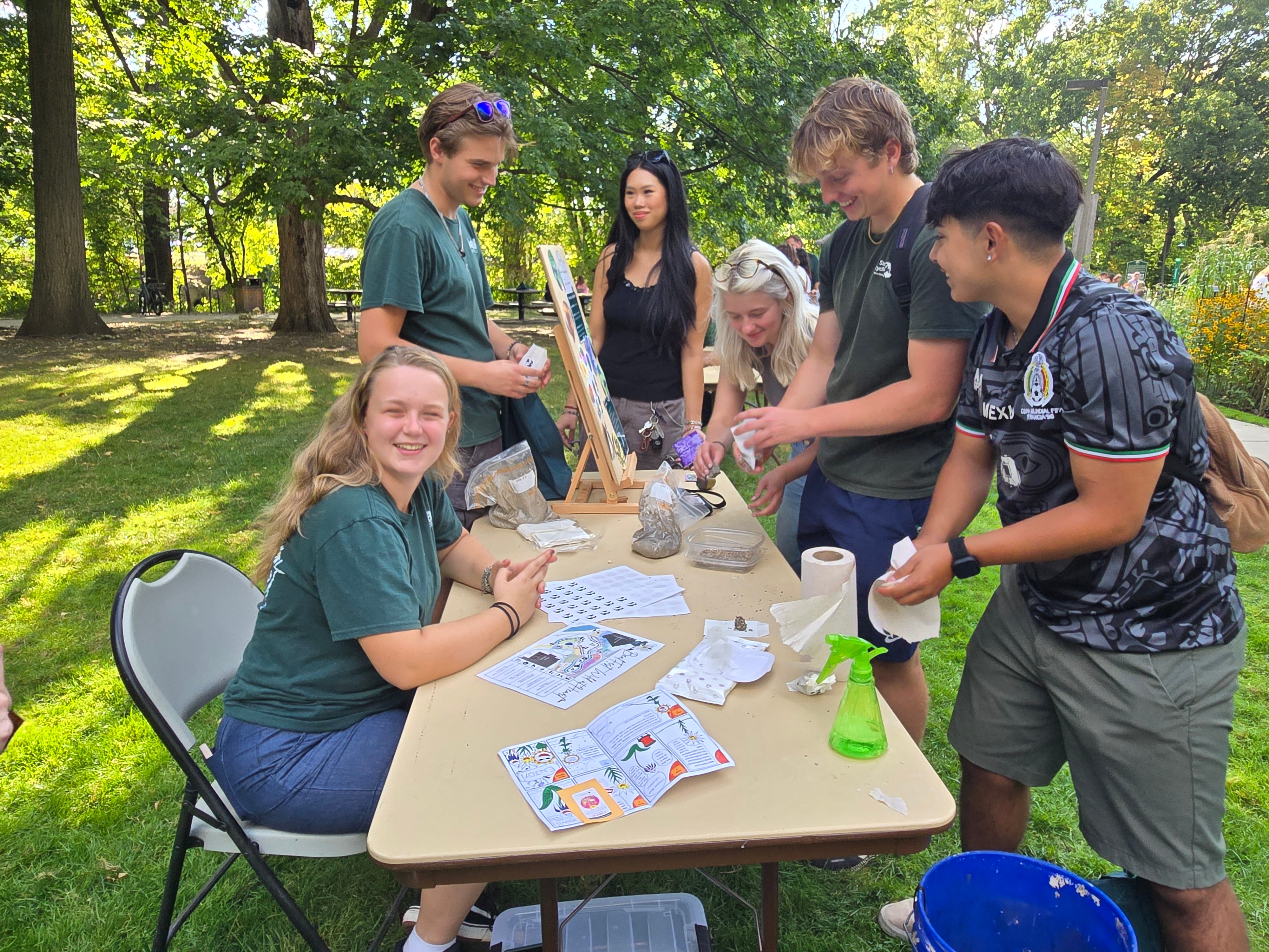 group of college students of various ethnicities and genders gathered around a table making a craft in a garden