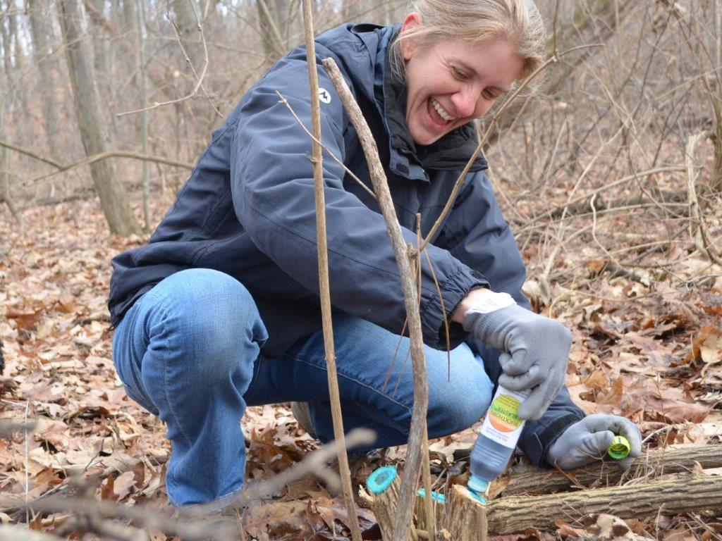 person wearing heavy winter clothing crouched in a forest with the ground covered in dead leaves holding a small bottle