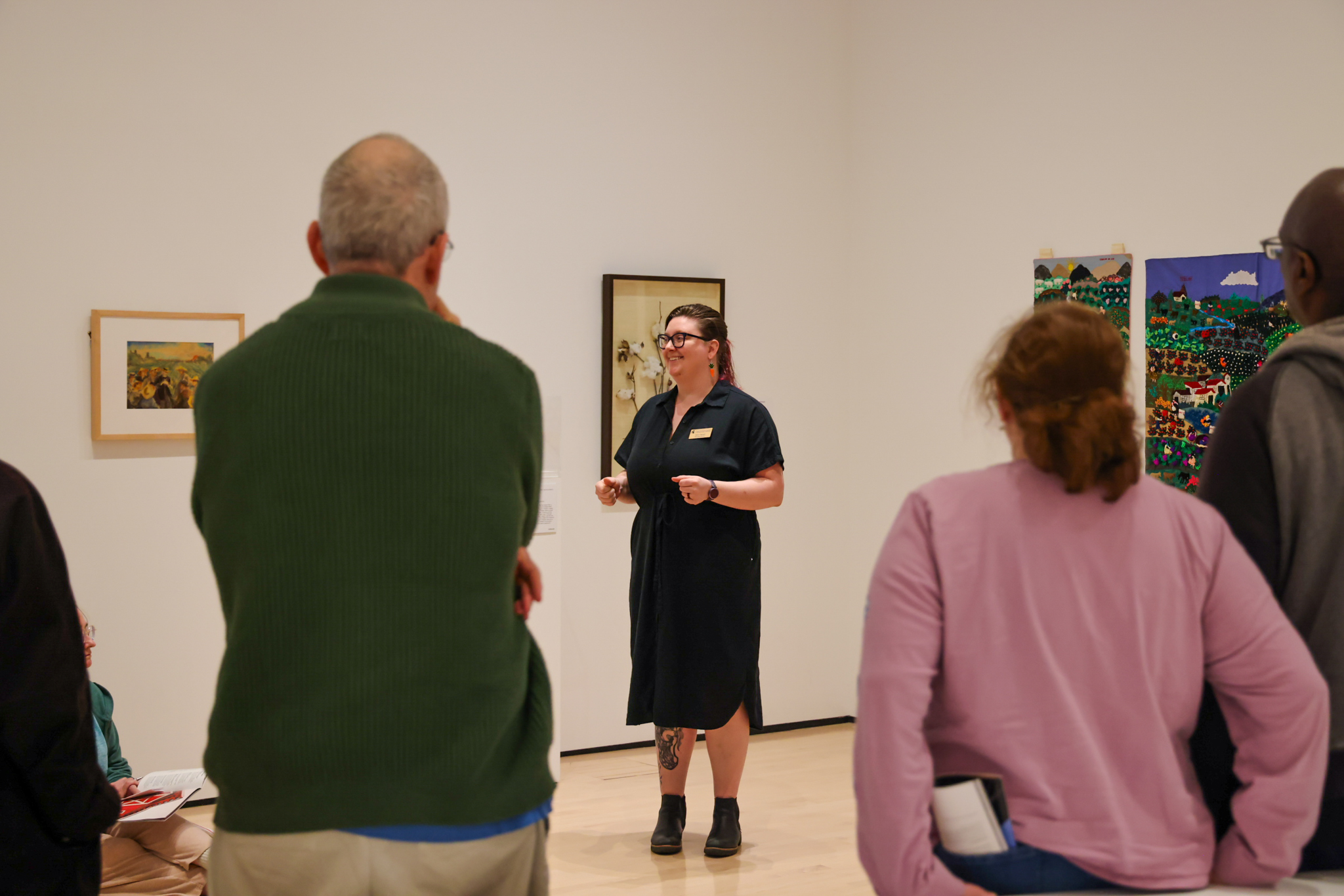 a woman with fair skin and dark hair pulled back in a ponytail wearing a black dress and dark rimmed glasses standing giving a tour in an art museum