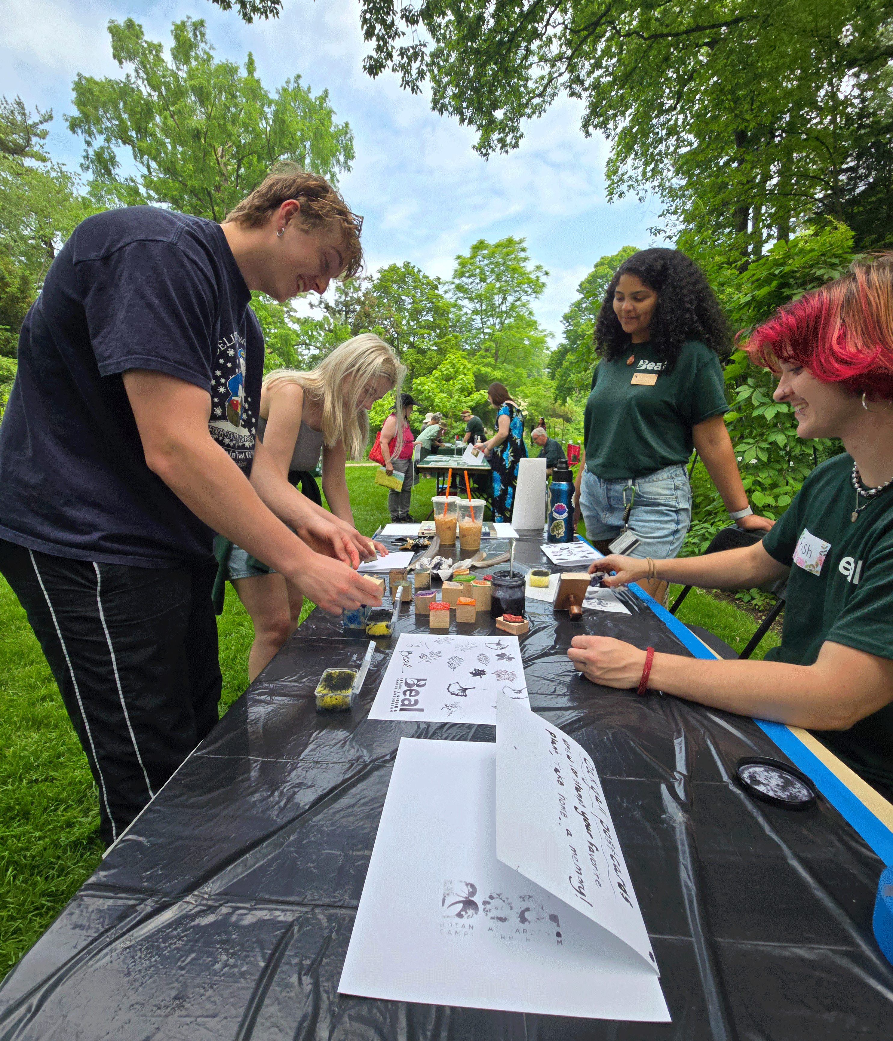 four people standing around a rectangular table doing an ink printing activity outside in a garden surrounded by trees
