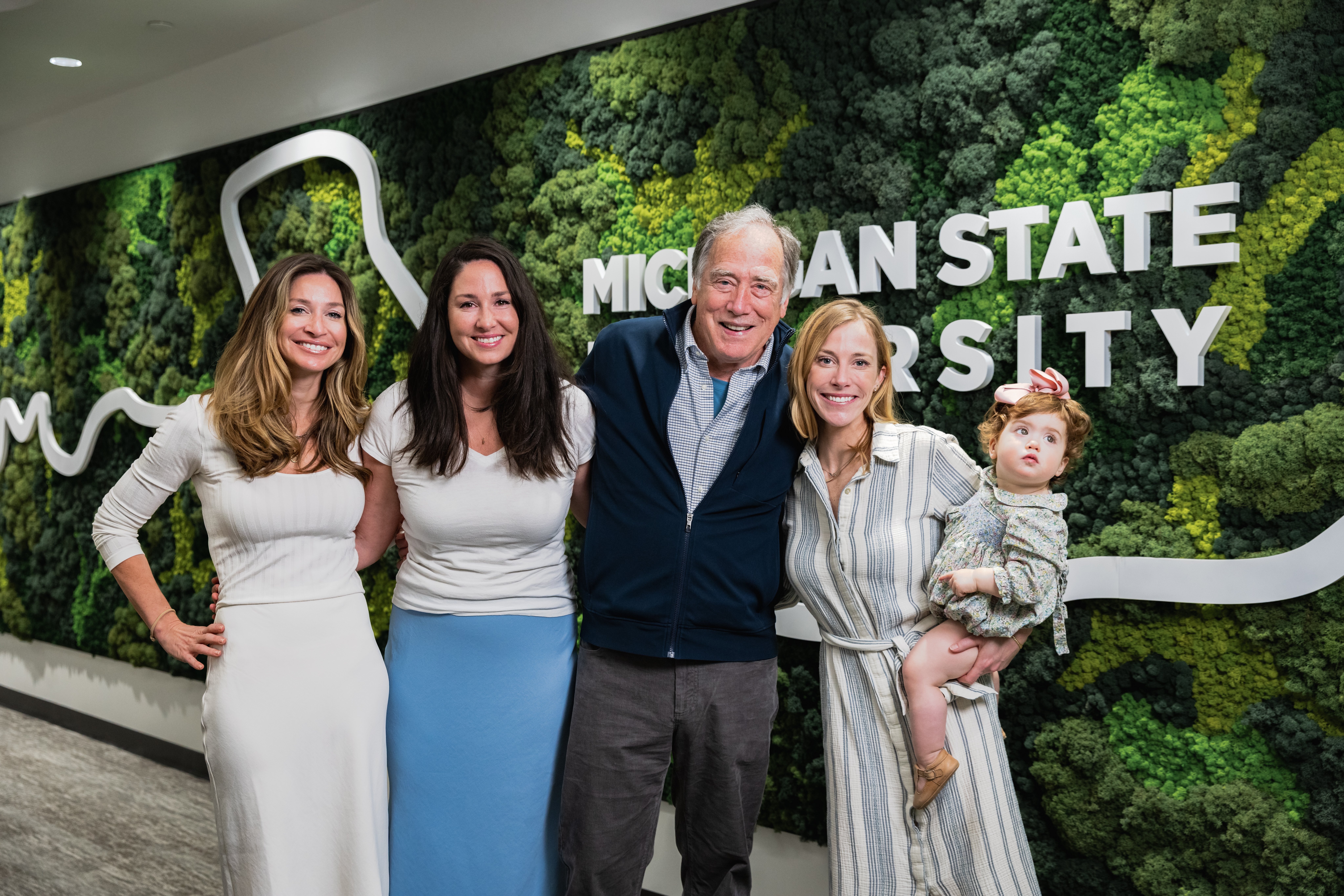 family photo of three adult women, one holding a toddler, and an older adult man all wearing buisness professional clothing standing in a hallway with a Michigan State University aerial photograph mural on the wall behind them