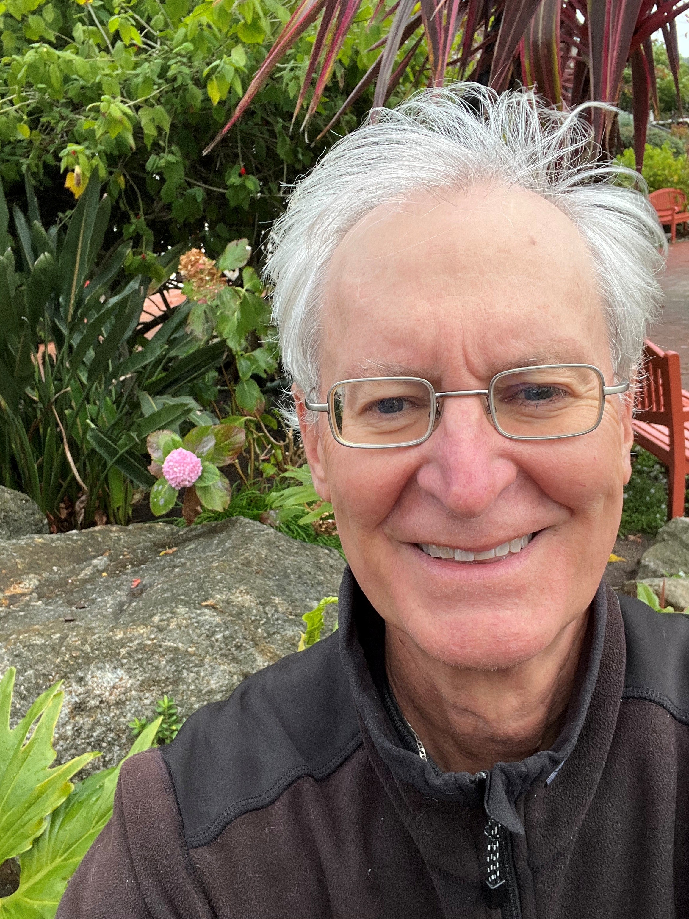 man with fair skin and gray hair wearing a dark winter coat and silver wire-rim glasses taking a smiling selfie in front of a green garden