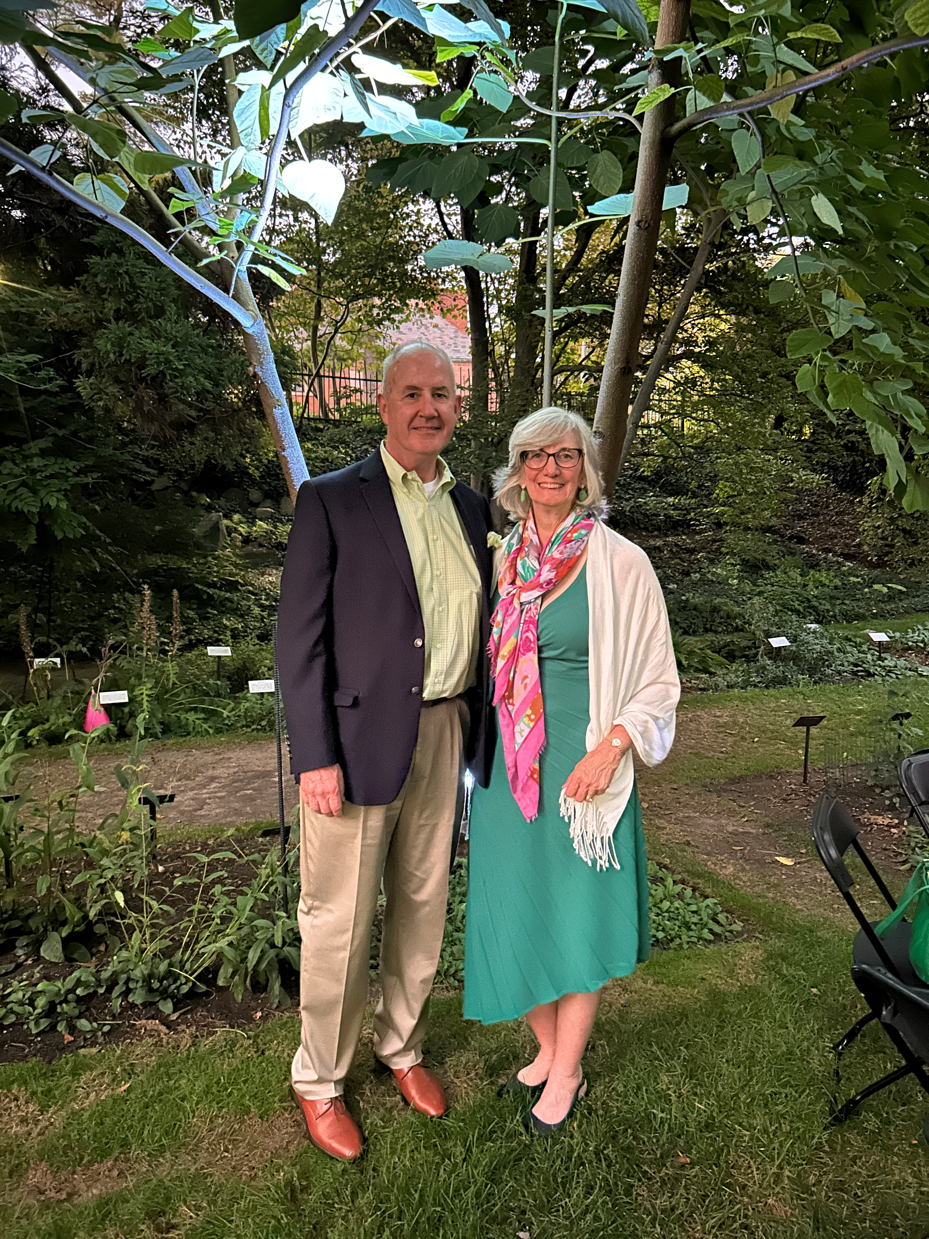 a man and a woman with grey hair wearing formal clothes posing together in a garden during an event