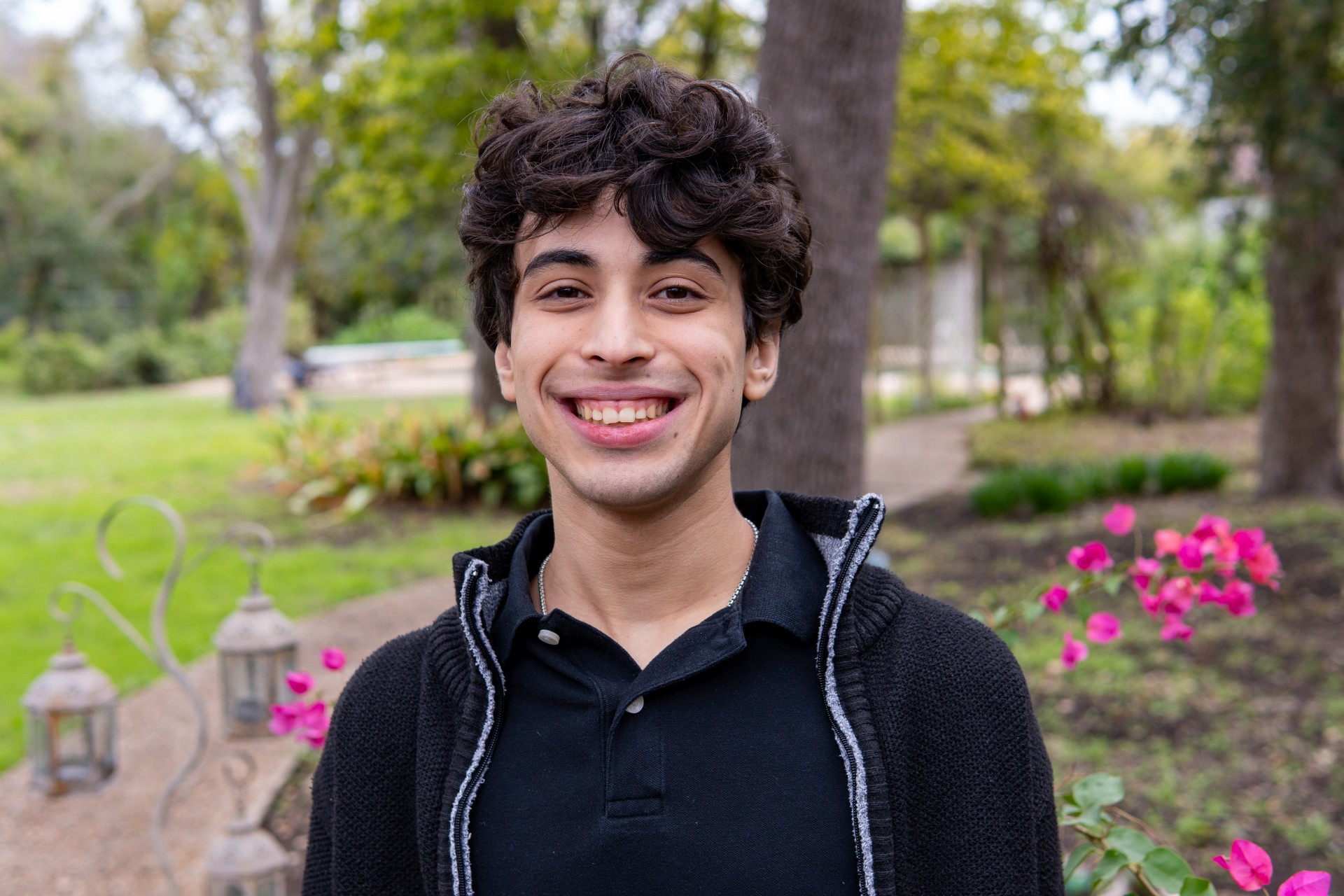 college aged man with curly dark hair and fair skin wearing a black sweatshirt smiling at a camera standing in a green garden with pink flowers behind him