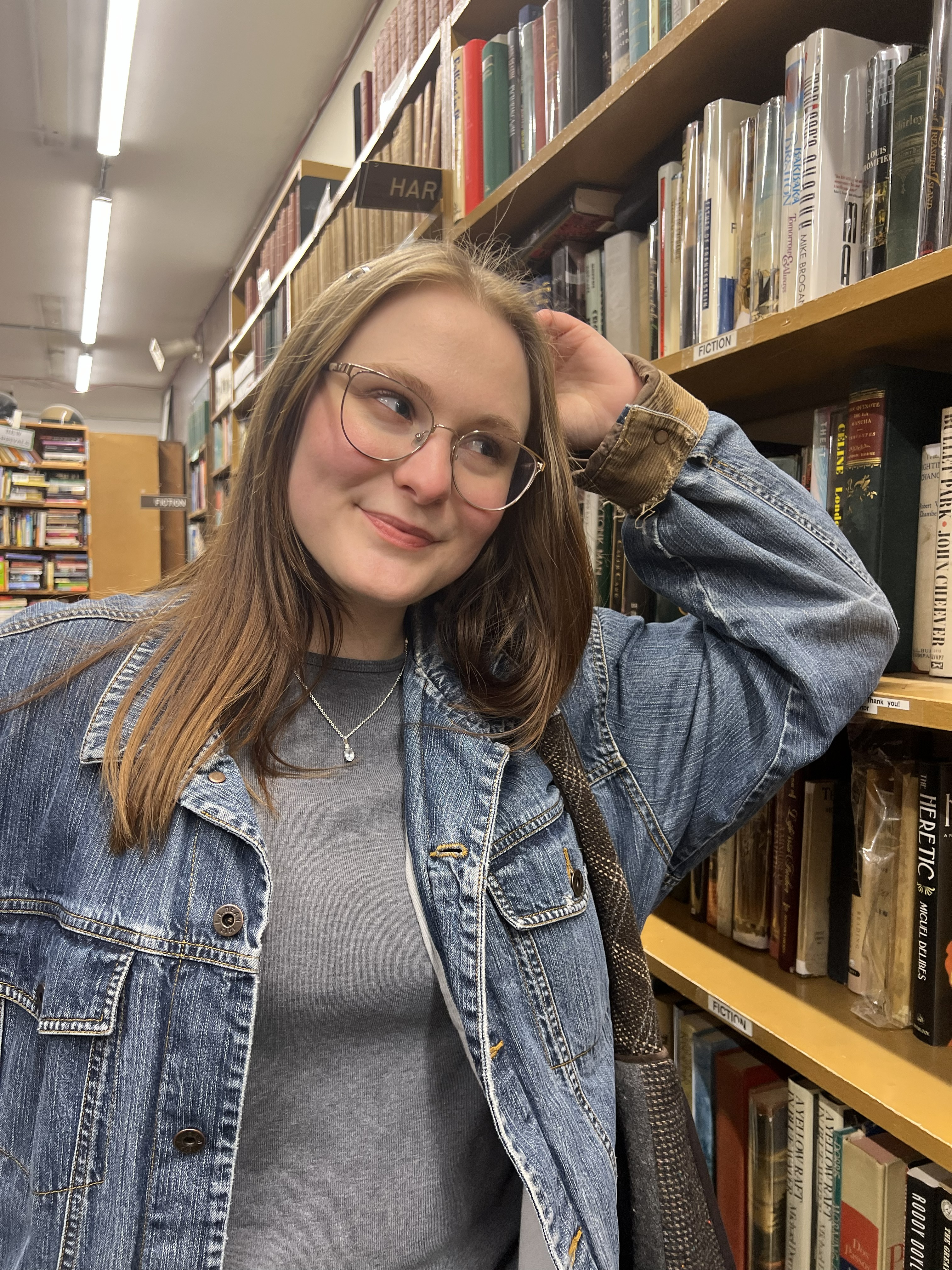 college aged woman with long blond hair and fair skin wearing a grey shirt and denim jacket and silver framed glasses standing leaning against a library bookshelf