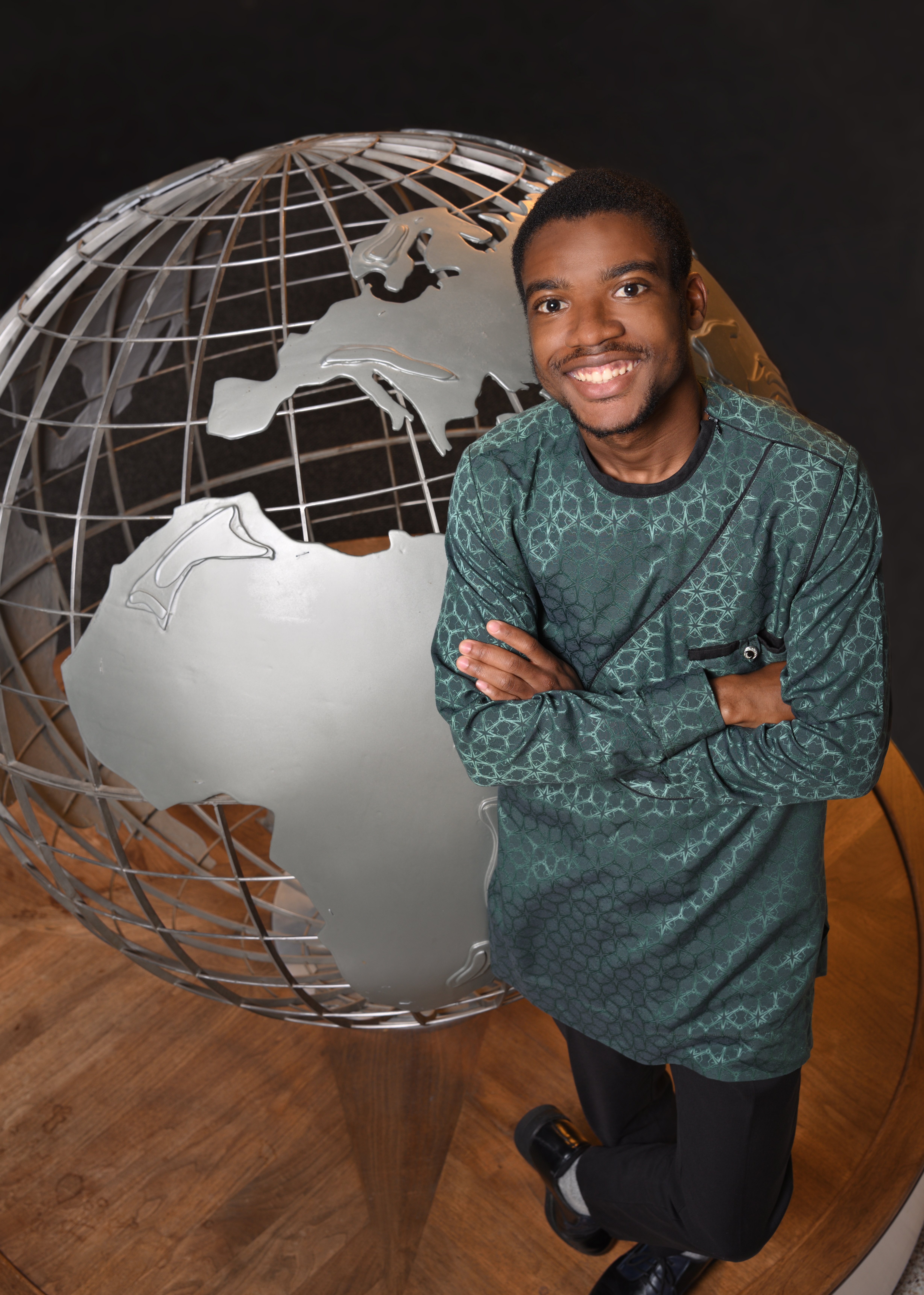 college aged man with dark skin and short dark hair wearing a green patterned shirt and black pants standing with arms crossed and smiling next to a large metal globe