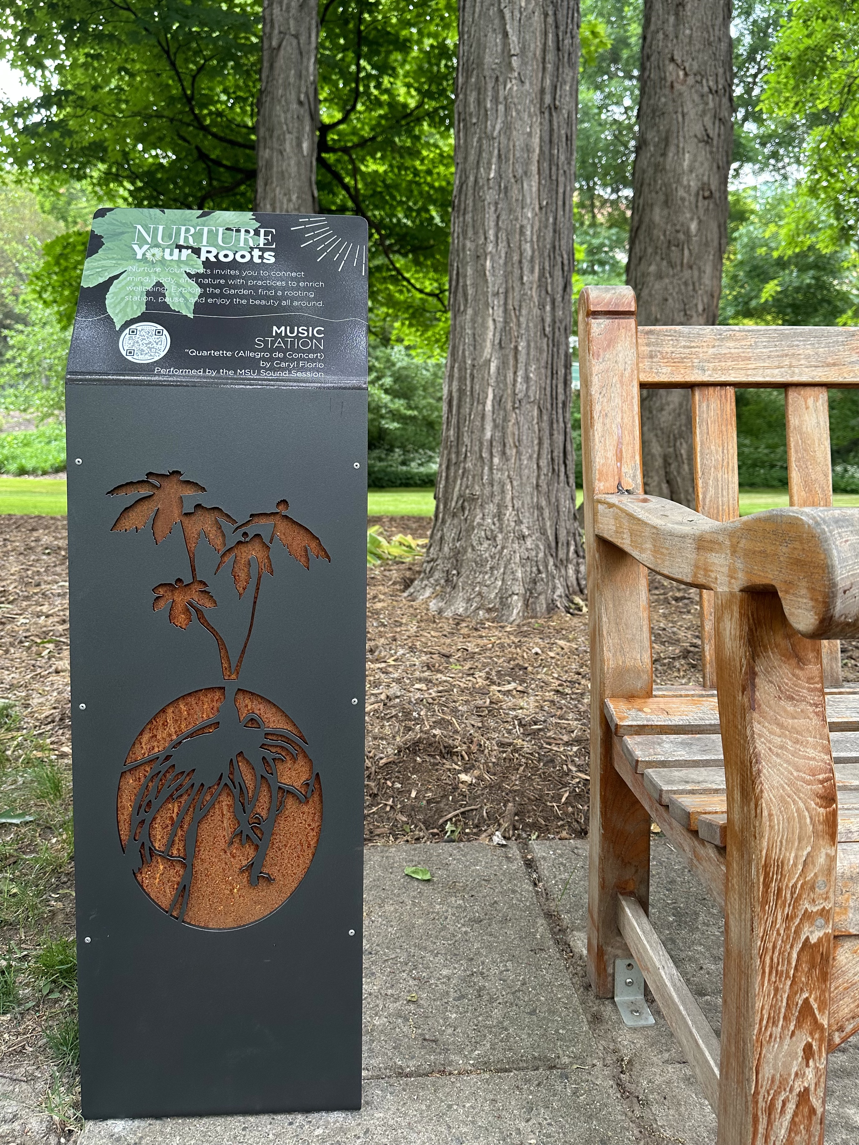 dark metal podium with Nurture Your Roots cutout logo of plant with roots next to a wooden bench with trees behind