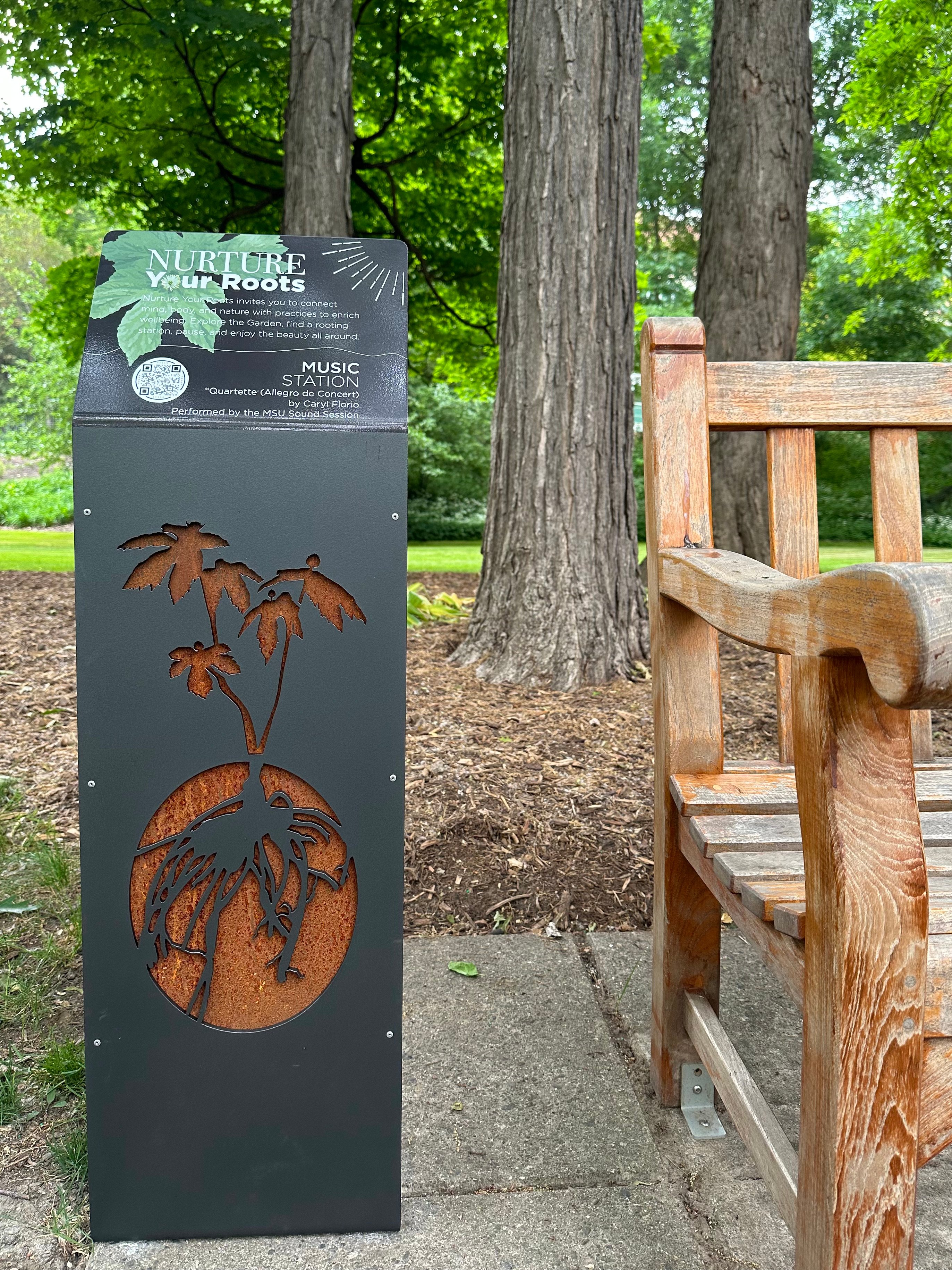 dark metal podium with Nurture Your Roots cutout logo of plant with roots next to a wooden bench with trees behind