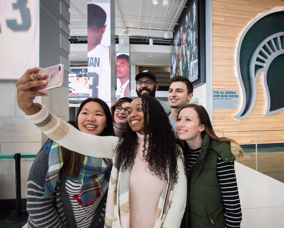 Group of diverse students taking a selfie indoors with a Spartan helmet in the background.