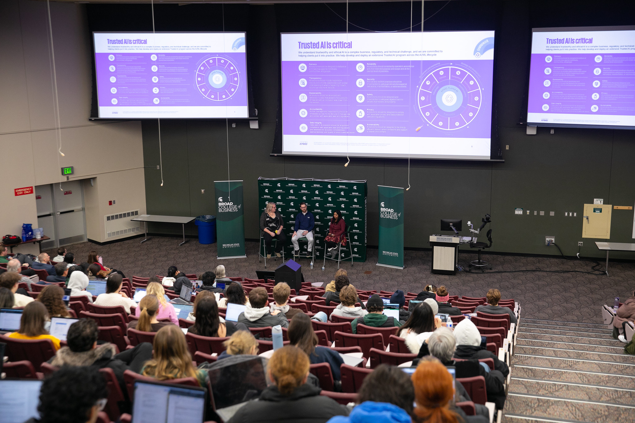 Three speakers sit beneath screens in a lecture hall and discuss artificial intelligence during an Ethics Week 2025 event.