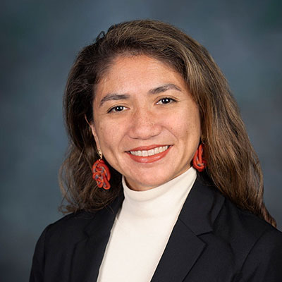 Headshot of Alessandra Carreon smiling, wearing red earrings, a white turtleneck, and black jacket, in front of a gray background.