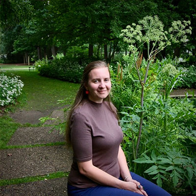 Katie Fry, smiling, wearing a brown shirt and blue jeans, sitting outside surrounded by green plants, flowers, and trees. 