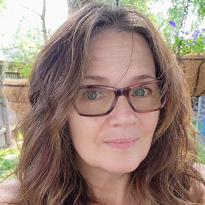 Headshot of Carla Iansiti, smiling, wearing glasses and a bright shirt, sitting outside with flower baskets in the background.