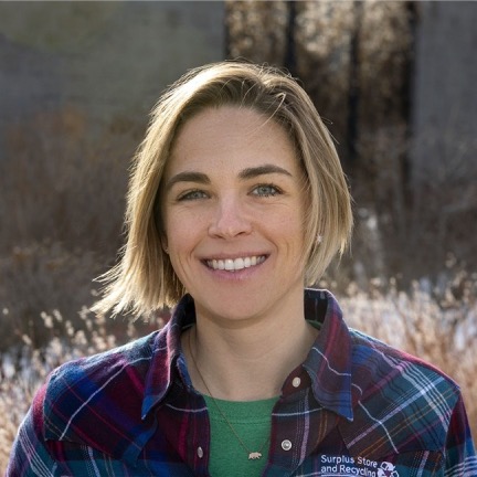 Katie Deska Radigan, smiling, wearing a green shirt and plaid jacket, standing outside in a field with trees in the background.