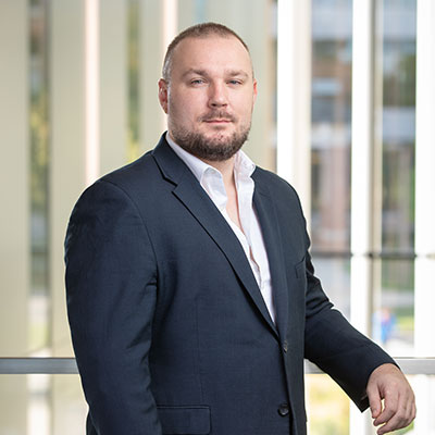 Headshot of Elliot Smith, wearing a dark suit and white shirt, standing in front of a glass railing in an office building.