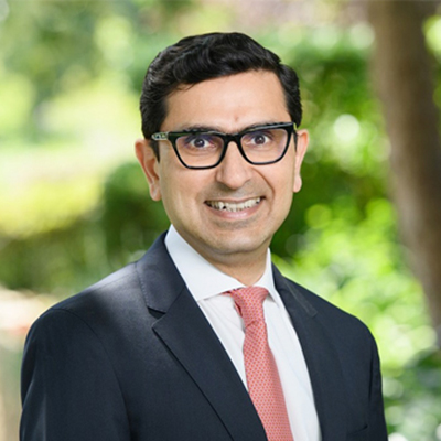 Headshot of Sarvesh Suri, smiling, wearing glasses and a dark suit with a pink tie, standing in front of trees.