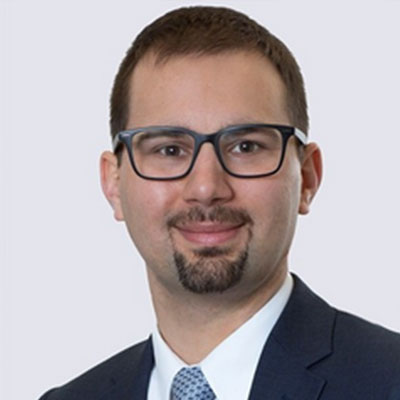 Headshot of Keith Underkoffler, smiling, wearing glasses and a dark suit with a white shirt and blue tie, standing in front of a white background.