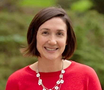 Courtney Venker, smiling, wearing a red blouse and a necklace, standing in front of greenery.