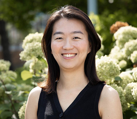 Kune Park, smiling, wearing a sleeveless black blouse and standing in front of blooming flowers.