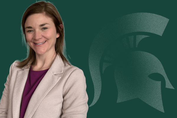 Nicki Schmidtke smiling, wearing a beige jacket with a cranberry blouse in front of a green background with a Spartan helmet logo.
