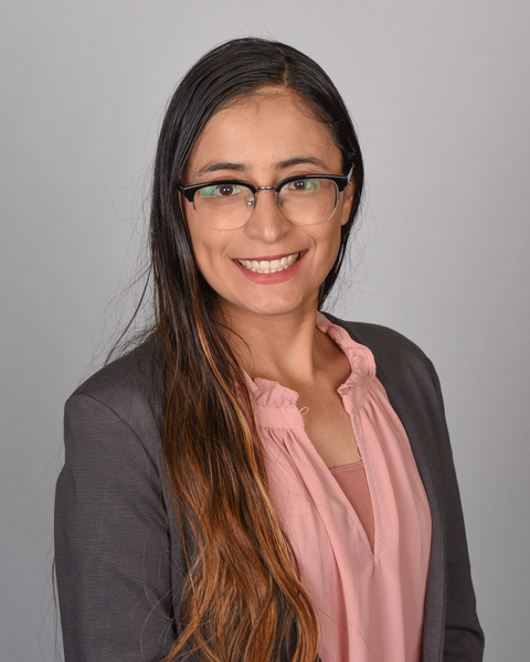 Connie Rojas in a pink shirt and grey overcoat, standing in front of a grey background.