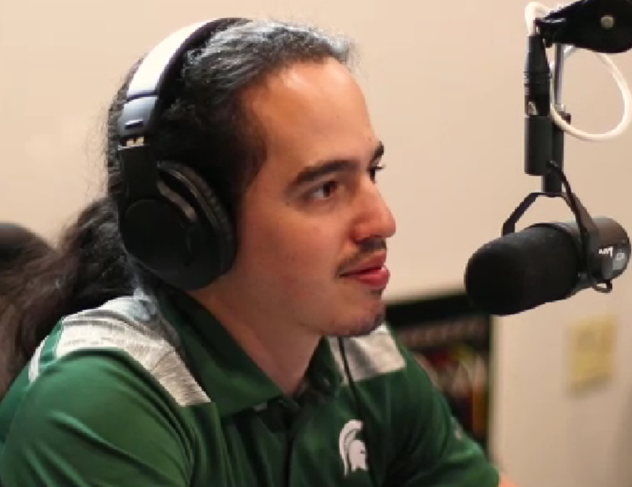 Daniel Puentes in an MSU shirt, wearing headphones and seated in front of a microphone.