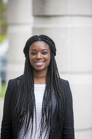 Courtney Bryant, dressed in a white shirt and black overcoat, standing in front of a pillar.
