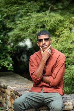 Ajamu Amiri Dillahunt dressed in an orange button up shirt, seated outside on a rocky ledge.