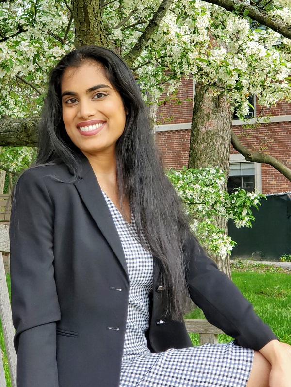 Chelsie Boodoo in a black suit coat and black and white checkered dress, sitting outside in front of a flowering tree.