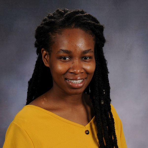 Gloria dressed in a yellow shirt, smiling in front of a grey background.