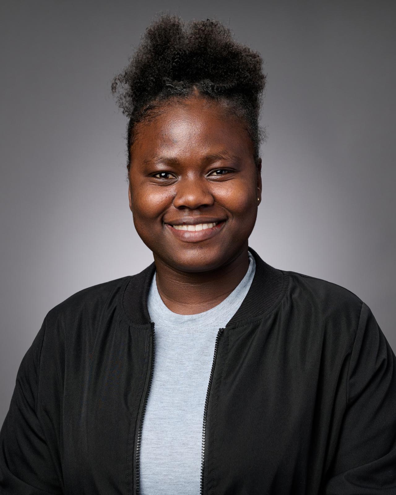 Catherine Baffloe smiles for a professional headshot against a gray background, wearing a black jacket over a light gray shirt.