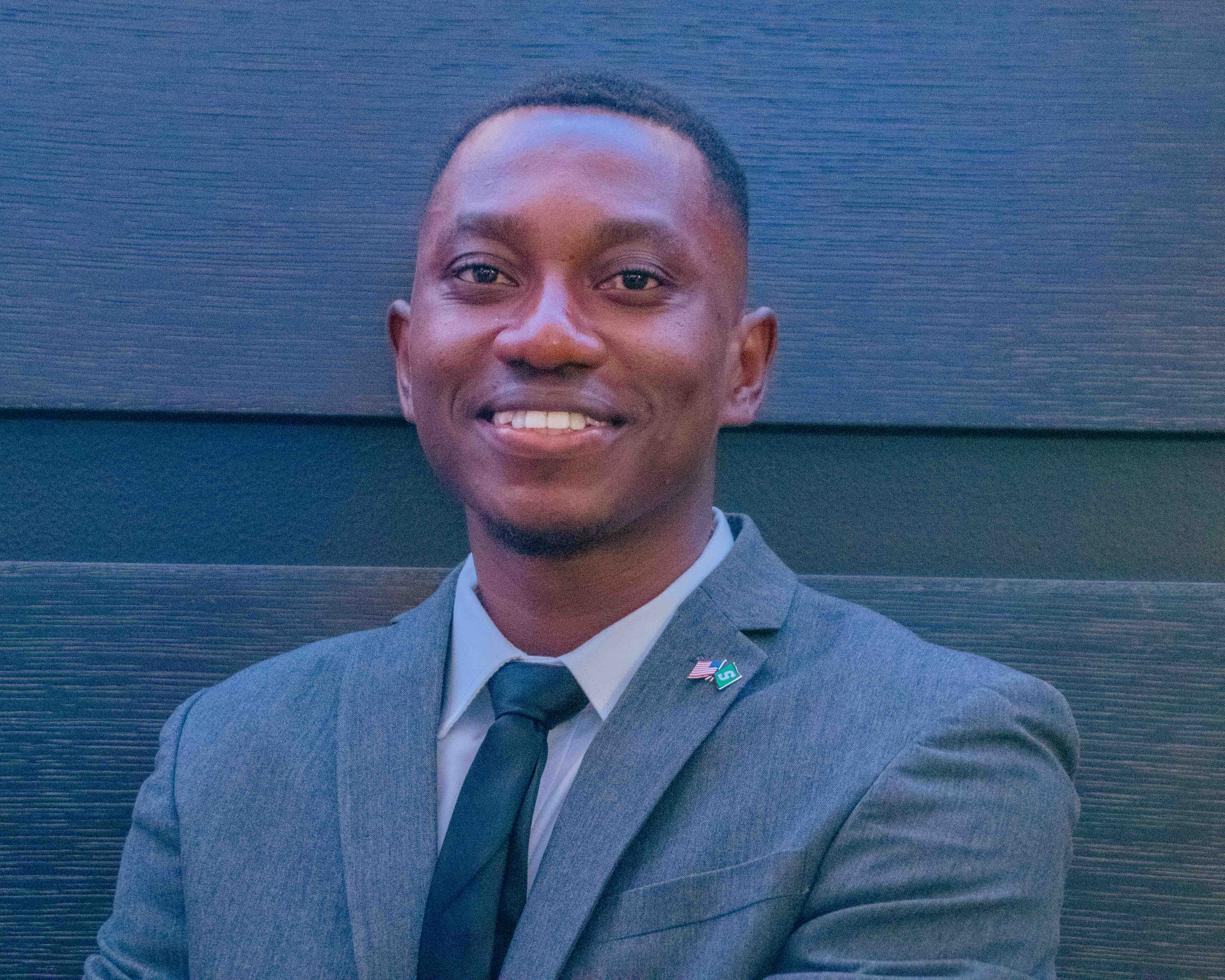 Samuel Ofosu smiles for a professional portrait, wearing a gray suit, light shirt, and dark tie with American and Michigan State University lapel pins.